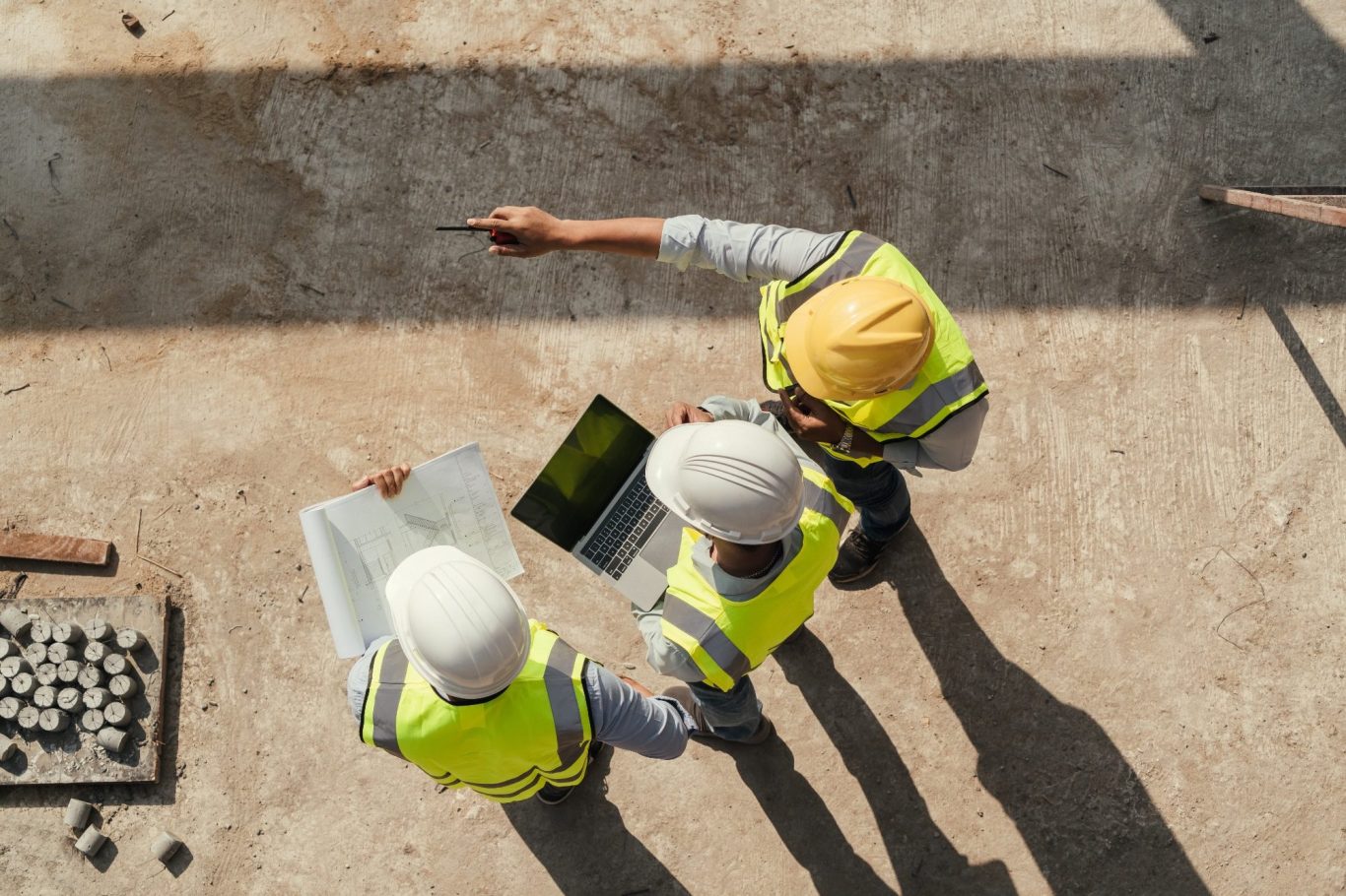 Drei Bauarbeiter mit Helmen prüfen Pläne und einen Laptop auf einer Baustelle.